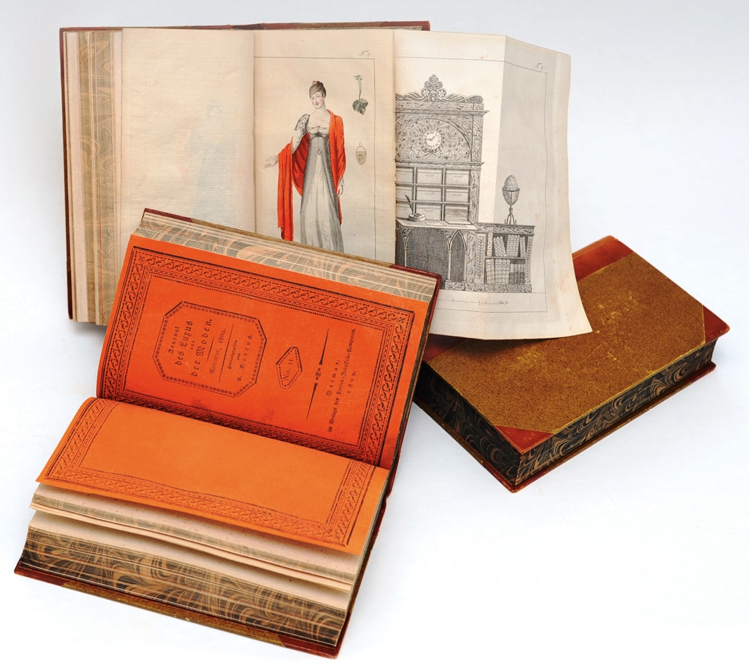 Verschiedene Bände des „Journals des Luxus und der Moden“. Aufgeschlagen: der orangefarbene Umschlag der Einzelhefte sowie Abbildungen einer Dame mit Samt-Tunika und eines Pariser Bibliothekssekretärs aus Mahagoni mit Verzierungen aus Bronze, Februar 1809;  Goethe-Museum Düsseldorf/Anton-und-Katharina-Kippenberg-Stiftung, Foto: Barbara Steingießer